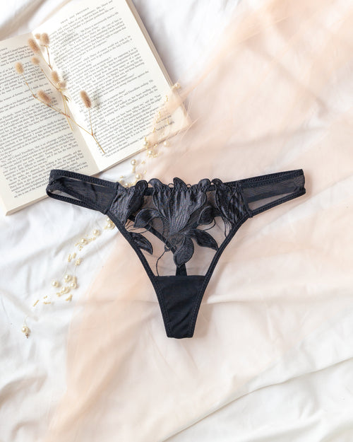 Black lace thong underwear laid flat on white fabric with an open book and dried flowers as styling props.