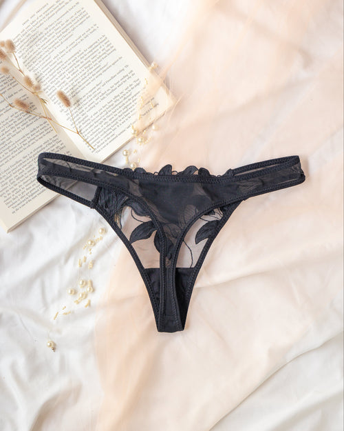 Black lace thong underwear laid flat on white fabric with an open book and dried flowers as styling props.
